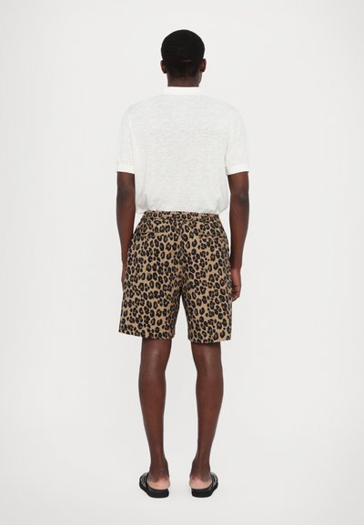 Leopard print shorts in tan with black spots, featuring a relaxed fit. Back view shows elastic waistband and pockets, worn with a light shirt.