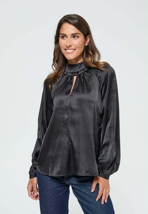 Blouse en satin noir avec col montant, détail froncé et découpe en forme de goutte. Manches longues avec poignets boutonnés. Portée avec un jean bleu foncé.