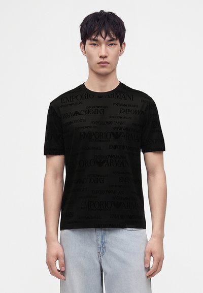 Νεαρός άνδρας με μαύρο T-shirt με το λογότυπο Emporio Armani και ανοιχτό μπλε τζιν, στέκεται μπροστά σε ένα απλό λευκό φόντο.