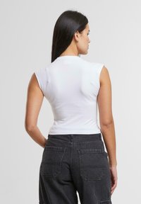 Weißes enges ärmelloses Oberteil mit hohem Kragen, getragen mit dunkelgrauen, hochgeschnittenen Hosen, die zwei Gesäßtaschen und einen strukturierten Stoff aufweisen.