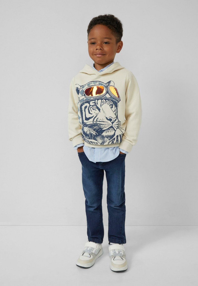 Crème hoodie met een blauwe tijgerafbeelding en oranje zonnebril, gelaagd over een blauw-wit gestreept shirt, gecombineerd met donkere jeans en sneakers.