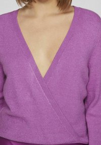 Fialový pletený svetr s hlubokým výstřihem do V a zavinovacím designem. Jemná textura a žebrované okraje zdůrazňují jeho strukturu.