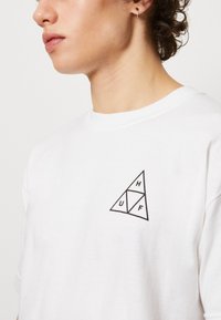 Camiseta de algodón blanca con un gráfico triangular negro que presenta las letras H, U y F. Mangas cortas y un cuello redondo. Ajuste informal.