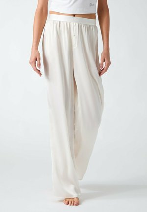 Mujer con pantalones blancos anchos y sueltos y una camiseta corta blanca, caminando descalza sobre un fondo blanco liso.