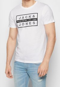 White cotton t-shirt featuring a black block graphic with text "JACK & JONES" and additional branding in contrasting fonts. Fitted design.