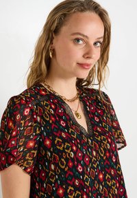 Blouse à manches courtes, à motifs en noir, rouge, vert et jaune ; possède un col en V avec une bordure en dentelle et un design ample et fluide.