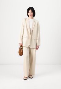 Krémově zbarvený oversized blazer se širokými kalhotami ve shodné barvě. Hladká tkanina, uvolněný střih, detailní kapsy. Drží malou hnědou kabelku.