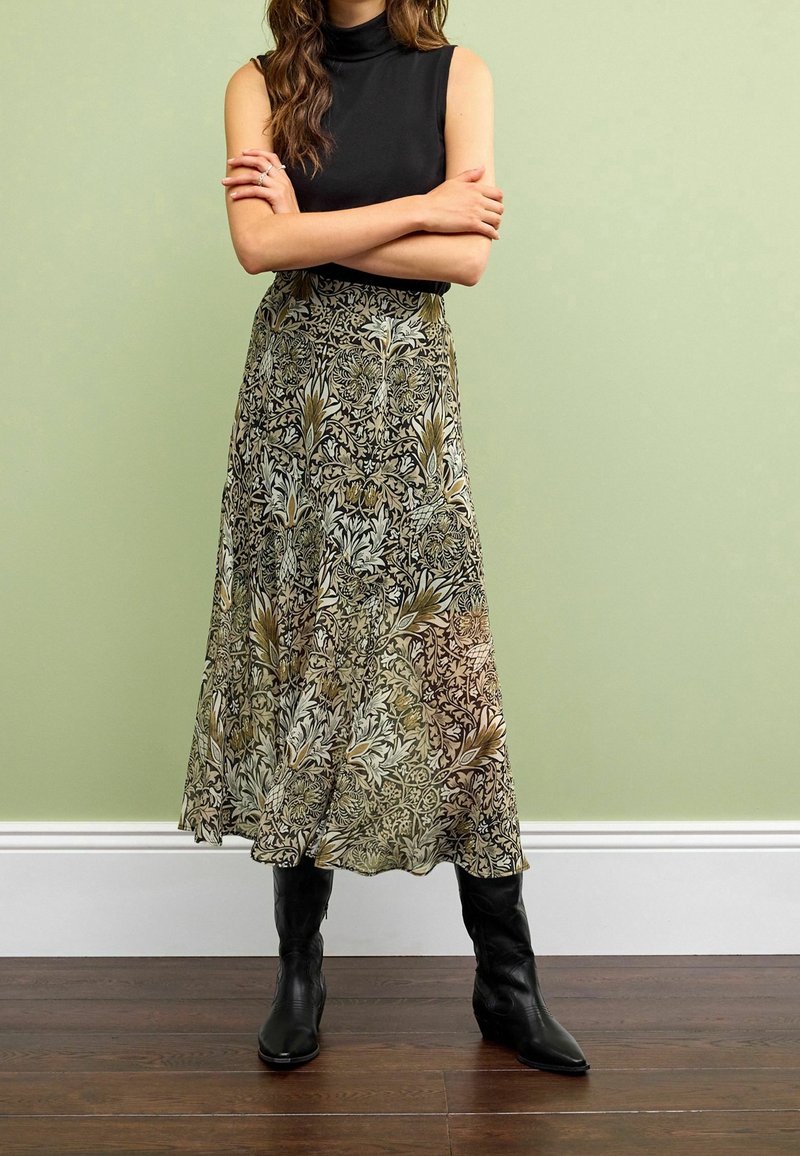 Femme portant un haut noir sans manches, une longue jupe à motif floral et des bottes noires, debout avec les bras croisés devant un mur vert clair.