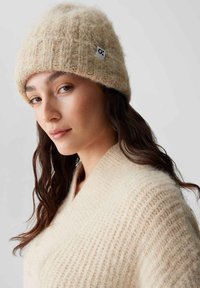 Berretto di lana beige a maglia con texture a coste e piccolo tag con logo; abbinato a un morbido maglione oversize color crema.