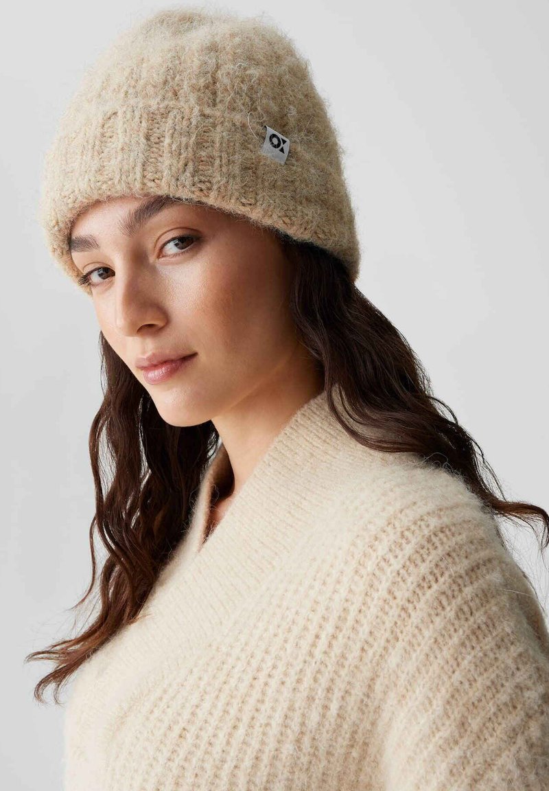 Berretto di lana beige a maglia con texture a coste e piccolo tag con logo; abbinato a un morbido maglione oversize color crema.