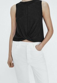 Haut sans manches noir à côtes avec un détail de torsion à l'avant, porté avec un pantalon blanc taille haute, une main dans la poche, sur un fond clair uni.