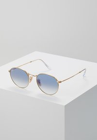 Occhiali da sole in metallo dorato con lenti rotonde a gradiente blu, aste sottili e terminali trasparenti. Design minimalista con un dettaglio di branding discreto.