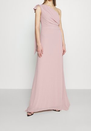 Donna che indossa un abito lungo fino al pavimento, rosa pallido, monospalla con corpetto drappeggiato e dettaglio a fiocco sulla spalla, abbinato a tacchi neri open-toe.