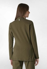 Oliivivihreä pitkähihaine takki, jossa on säädettävä istuvuus, näkyvät tikkausdetailit ja pieni lappu takana. Pehmeä, tekstuurinen kangas.