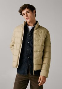 Chaqueta acolchada beige con diseño de acolchado vertical, cierre de cremallera en la parte delantera y cuello alto, llevada sobre una camisa de mezclilla oscura. Textura suave, ligera.
