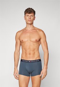 Männerboxershorts aus einem einfarbigen dunkelblauen Stoff mit einem schwarzen Bund, der das Logo "TOMMY HILFIGER" in Weiß trägt. Glatte Textur, figurnahes Design.
