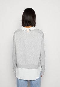Sweatshirt gris avec une chemise à col blanc par-dessus. Présente une ouverture en forme de trou de serrure à l'arrière et des poignets côtelés. Texture douce.