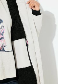Lichtgekleurde jas met zwarte fleece voering. Voorzien van een ritszak en contrasttexturen, die een mix van materialen en tinten laten zien.