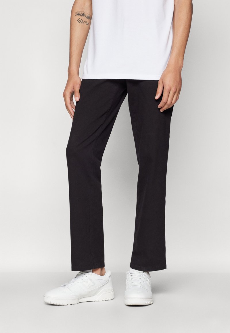 Personne portant un pantalon noir droit, des baskets blanches et une chemise blanche, debout devant un fond uni.
