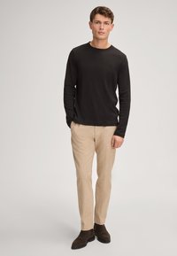 Schwarzes Langarmshirt, kombiniert mit hellbeigen Hosen. Das Model steht in dunklen Schuhen vor einem neutralen Hintergrund.