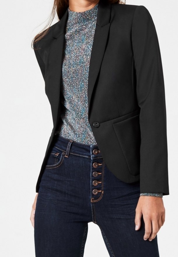 Femme portant un blazer noir ajusté sur un haut à col montant tacheté et un jean taille haute bleu foncé avec fermeture à boutons visible.