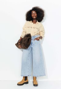 Chemisier en dentelle beige à manches longues, jeans larges taille haute bleu clair, bottines en daim tan à talon, et un grand sac en daim marron.