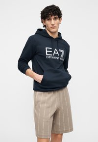 Tengerészkék kapucnis pulóver első zsebbel és húzózsinórral, fehér logós felirattal. Bézs rövidnadrággal párosítva, amelyen ismétlődő logóminta van domborítva.