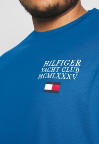 Modré bavlněné tričko s bílým textem "HILFIGER YACHT CLUB MCMLXXXV" a obdélníkovým logem v červené, bílé a černé barvě.