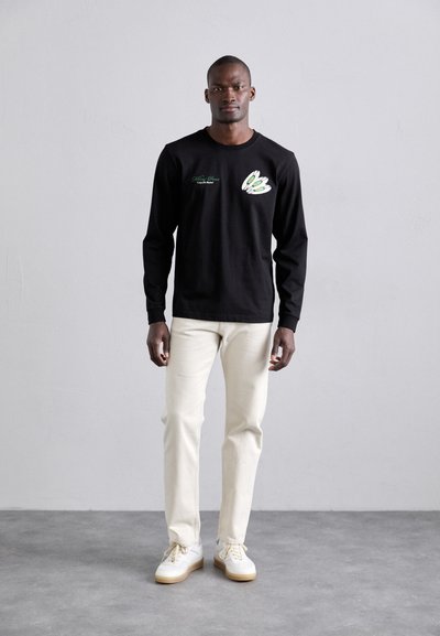 Black long-sleeve shirt with green graphic detail on the left chest, paired with cream-colored pants and white shoes with gum soles.