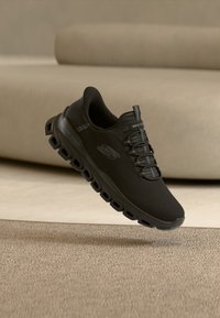 Zapatilla deportiva Skechers negra con diseño único de suela hueca, flotando sobre una alfombra beige y junto a un sofá beige.