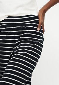 Άτομο που φοράει μαύρο παντελόνι με λευκές οριζόντιες ρίγες, με το χέρι στην τσέπη, σε συνδυασμό με λευκό πάνω μέρος.