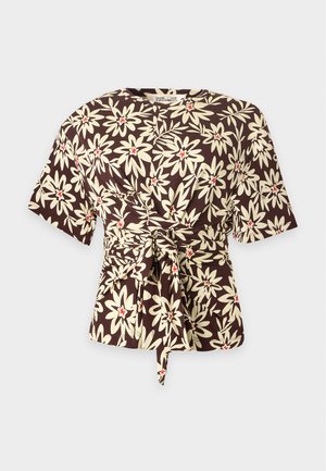 Femme portant un haut fleuri marron et une jupe blanche fendue haute avec des sandales noires à talons et un sac marron bandoulière, debout devant un fond uni.