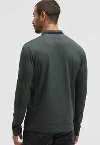 Camicia a maniche lunghe di colore verde scuro con colletto a girocollo, caratterizzata da una finitura testurizzata e un logo stampato sulla parte superiore della schiena e sull'orlo inferiore.