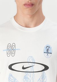 White cotton t-shirt featuring blue graphic prints, a black Nike logo, and various designs including circular shapes and icons.