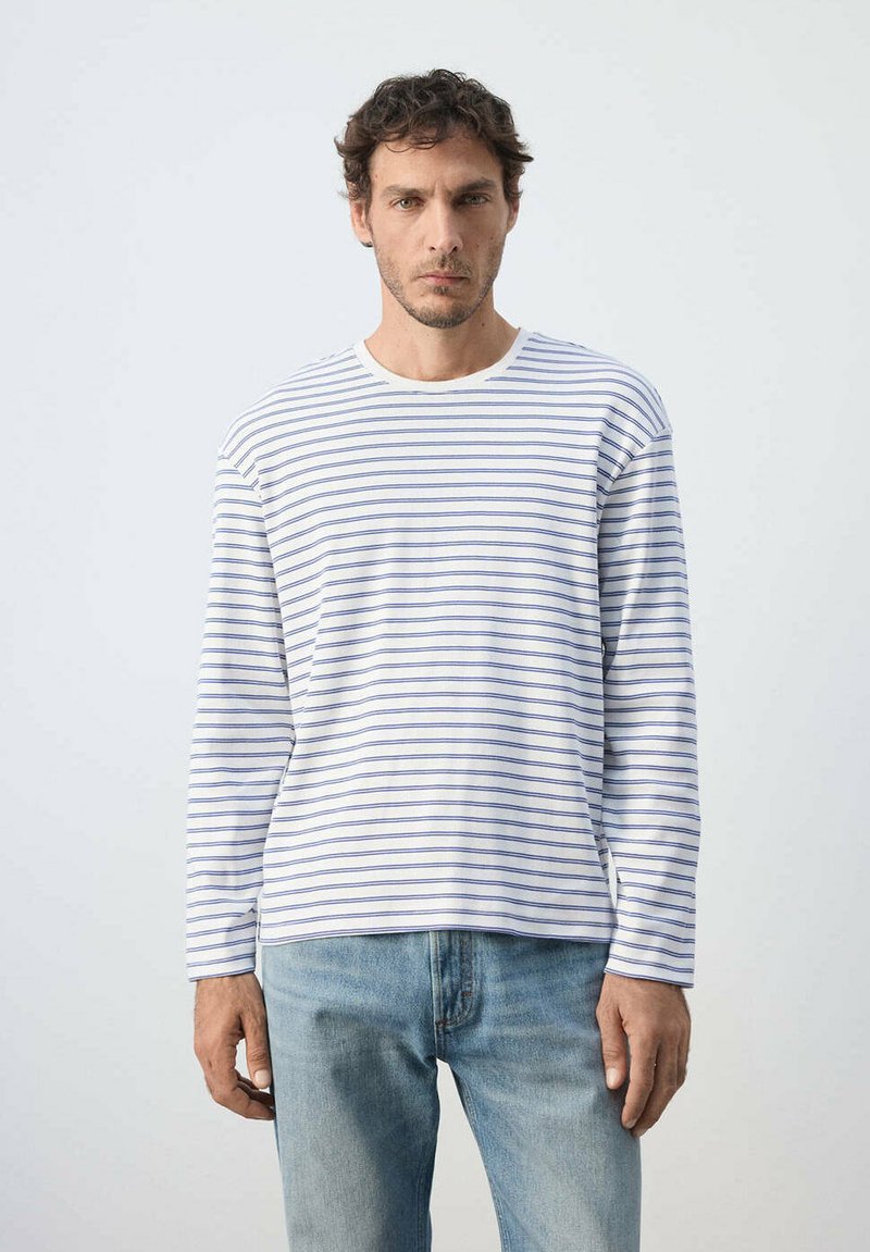Homme portant une chemise blanche à manches longues avec de fines rayures horizontales bleues et un jean bleu clair, debout devant un fond uni.