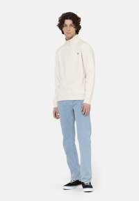 Sudadera cremosa con cierre y puños acanalados, combinada con jeans de mezclilla azul claros y zapatillas negras. Diseño simple con detalles mínimos.