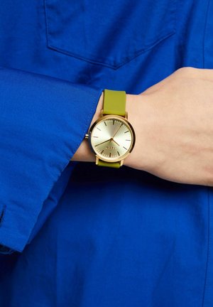Goldfarbene Uhr mit rundem Ziffernblatt und strukturiertem hellgoldenem Ziffernblatt. Grüner Silikonarmband, am Handgelenk getragen, vor einem blauen Hemd-Hintergrund.