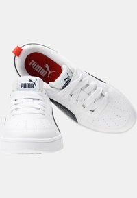 Sneaker bianche con accenti neri, dotate di una parte superiore testurizzata, punta tonda, suola piatta e dettagli perforati. Include un'etichetta con logo rosso e nero.