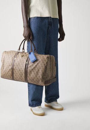 Borsa da viaggio a fitta beige con dettagli in pelle marrone, forma strutturata, manici doppi e etichetta per bagagli blu. Persona vestita con jeans blu e sneaker.
