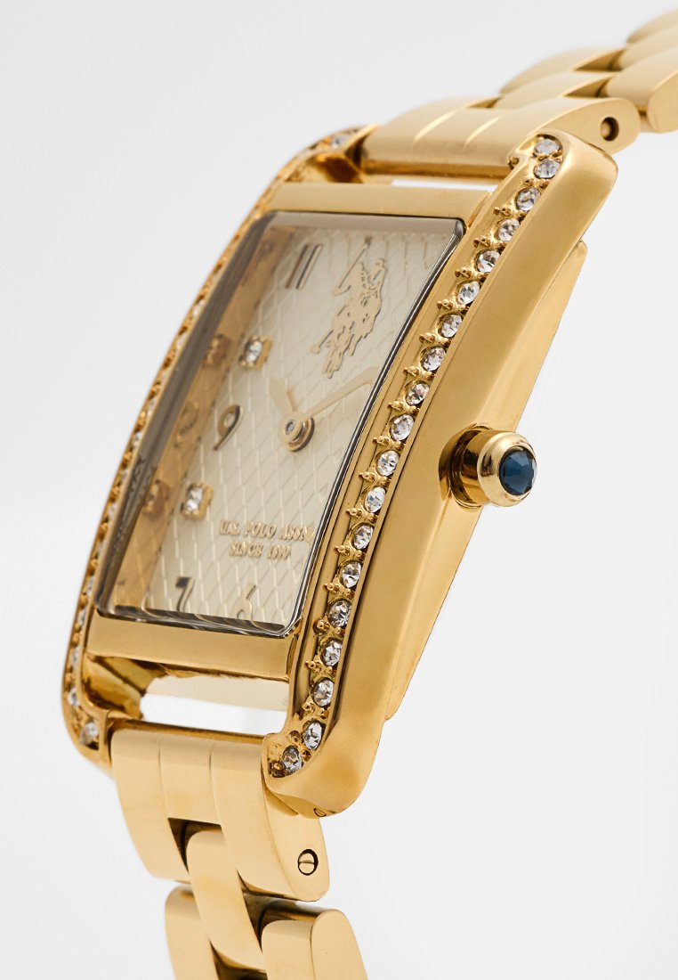 Goldfarbene rechteckige Armbanduhr mit einem strukturierten, hellgoldenen Zifferblatt, Strass-Akzenten und einem blauen Cabochon auf der Krone. Armband aus Metallgliedern.