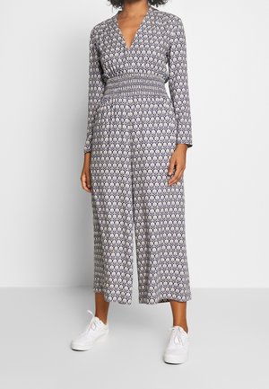 Combinaison à motifs navy avec des manches longues, décolleté en V, taille smockée et silhouette à jambes larges, associée à des baskets blanches.