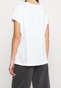 Personne portant un t-shirt blanc uni à manches courtes et un pantalon gris foncé, vue de dos sur un fond uni.