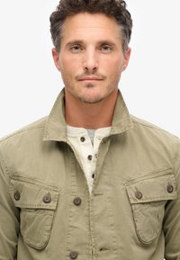 Khaki knoopjasje met twee borstzakken, voorzien van een kraag en houten knopen. Daaronder een lichtgrijs henley shirt met knopen.