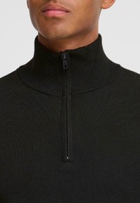 Gros plan sur le cou et le menton d'un homme portant un pull noir zippé et à col montant, sur un fond neutre.