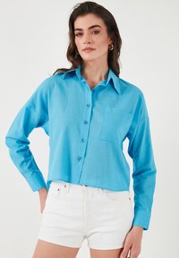 Light blue, cropped button-up shirt in a smooth fabric with long sleeves, single chest pocket, and matching blue buttons. Paired with white shorts.