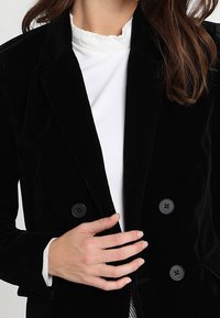 Černý sametový dvouřadý blejzr se šesti černými knoflíky, klopovými límci a přiléhavým střihem, navrstvený přes bílou blůzu.