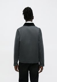 Mand med mørke dreadlocks iført en mørkegrå jakke og sorte bukser, stående med ryggen til mod en ensfarvet lys baggrund.