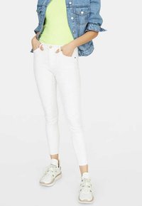 Jeans blancos ajustados de cintura alta, combinados con una chaqueta de mezclilla azul clara sobre una blusa verde lima. Zapatillas blancas con detalles en beige.