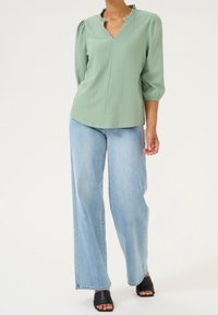 Blouse texturée verte avec col en V et col à volants, associée à un jean large bleu clair et des sandales noires ouvertes à enfiler.