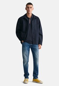 Chaqueta azul marino con cierre de botones y bolsillos, combinada con un polo negro con un cuello de tres rayas, vaqueros azules y zapatillas amarillas.